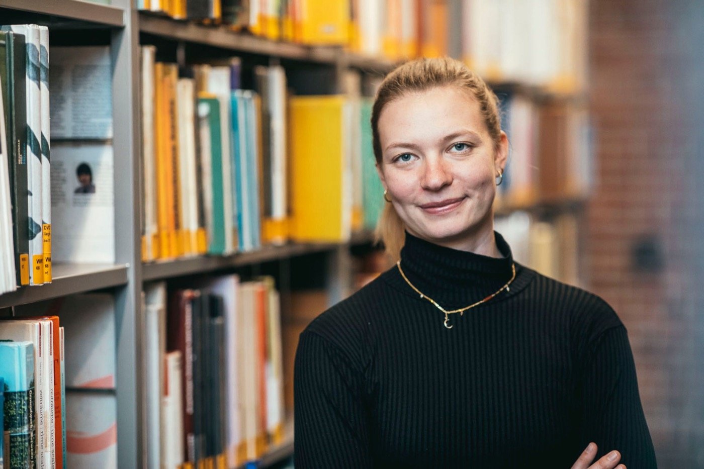 Stephi Bünemann steht mit verschränkten Armen vor einem Bücherregal und lächelt